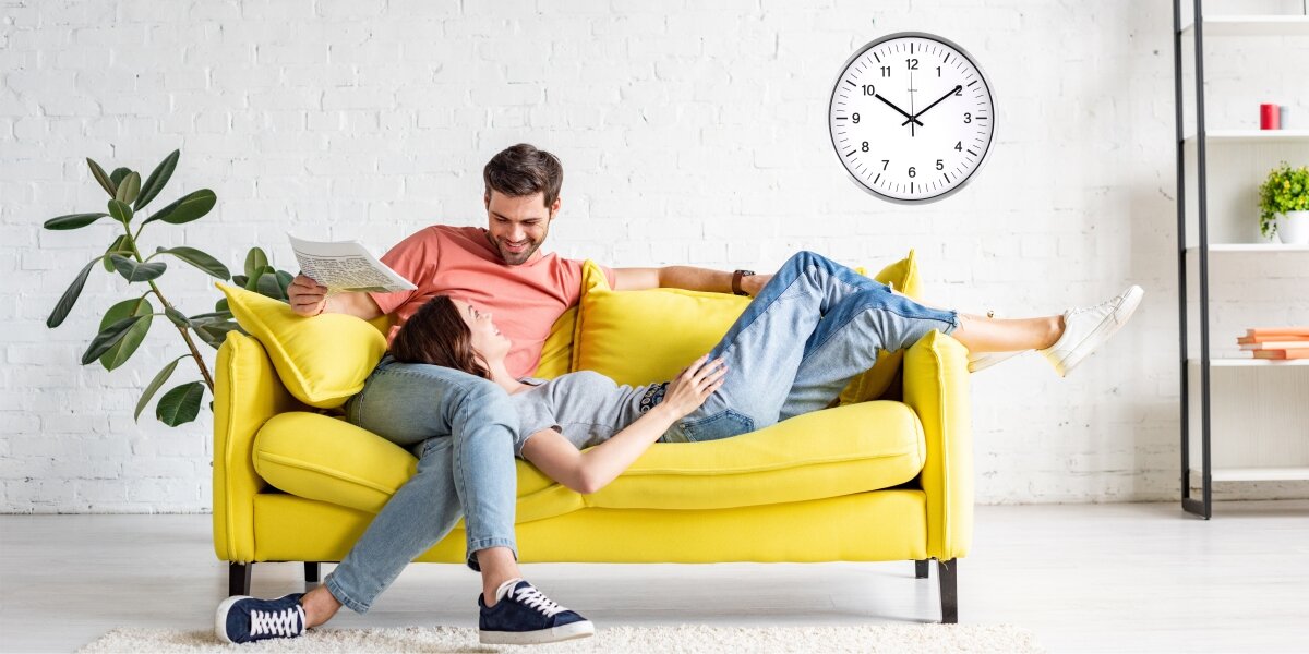 Zegar HAMA Java Srebrny, Czas pod ręką, Para spoczywa na razem na kanapie, w tle zegar na ścianie