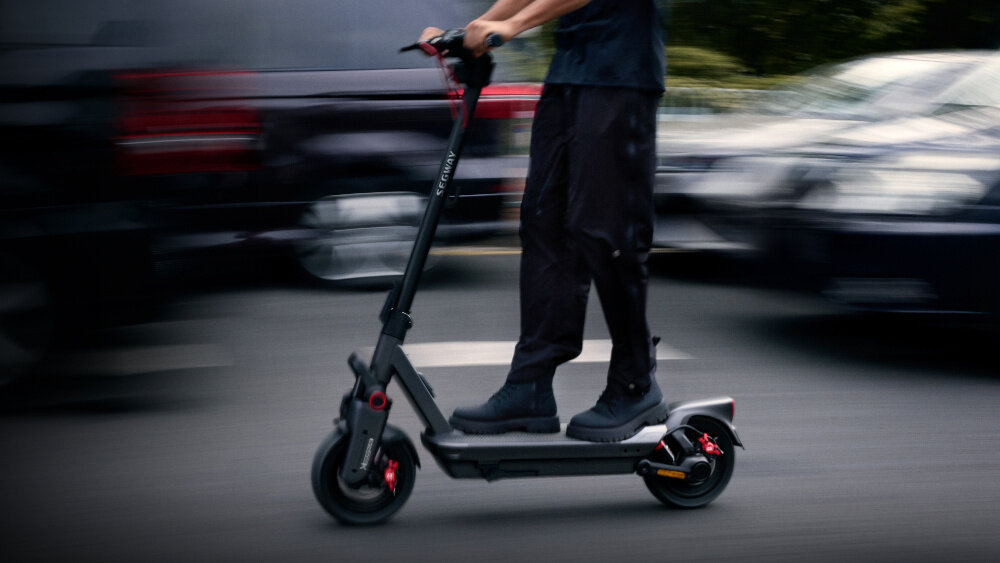 Hulajnoga elektryczna SEGWAY Ninebot Max G3 D 80km 2000W 2 amortyzatory 11 cali Czarny miasto ulica osoba jedzie na hulajnodze zbliżenie na podest w tle samochody wytrzymała konstrukcja z wysokiej jakości aluminium dla osób o maksymalnej wadze do 130 kg spory deck komfortowa i stabilna jazda antypoślizgowa powłoka