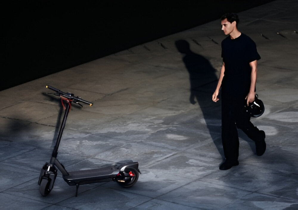 Hulajnoga elektryczna SEGWAY Ninebot Max G3 D 80km 2000W 2 amortyzatory 11 cali Czarny miasto chodnik osoba podchodzi do hulajnogi hulajnoga oparta na stopce stoi na chodniku składana stopka nóżka zamontowana w centralnym punkcie ciężkości sygnał dźwiękowy bezpieczeństwo