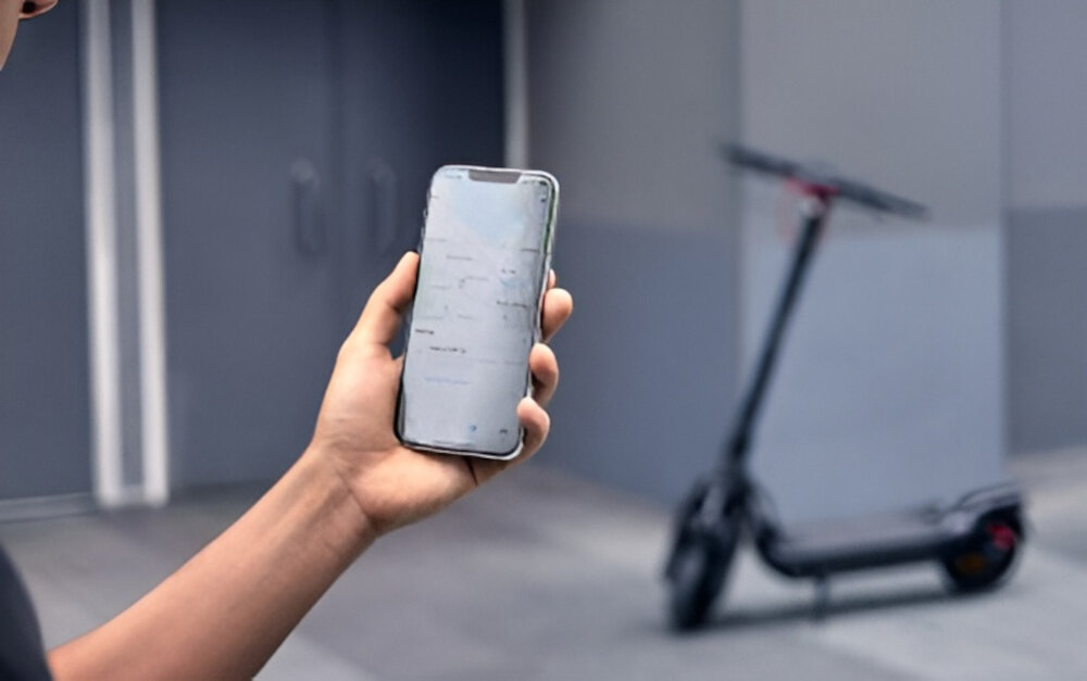 Hulajnoga elektryczna SEGWAY Ninebot Max G3 D 80km 2000W 2 amortyzatory 11 cali Czarny miasto osoba siedzi w budynku trzyma telefon w dłoni przed budynkiem stoi hulajnoga wbudowany moduł Bluetooth aplikacja mobilna Segway-Ninebot Smart App na systemy Android i iOS