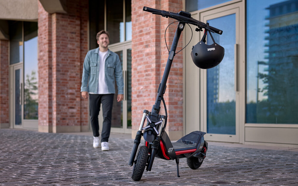 Hulajnoga elektryczna SEGWAY Ninebot ZT3 Pro D 70km 1600W Amortyzacja 11 cali Czarny miasto chodnik osoba podchodzi do hulajnogi hulajnoga oparta na stopce stoi na chodniku w tle budynek z dużymi oknami składana stopka nóżka zamontowana w centralnym punkcie ciężkości sygnał dźwiękowy bezpieczeństwo