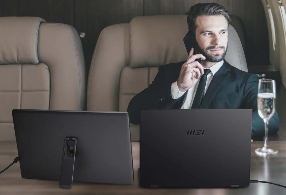 Monitor MSI Pro MP161 E2U mężczyzna w garniturze siedzi w samolocie z laptopem i dodatkowym ekranem, rozmawia przez telefon, ochrona wzroku, redukcja migotania, filtr niebieskiego światła, certyfikat