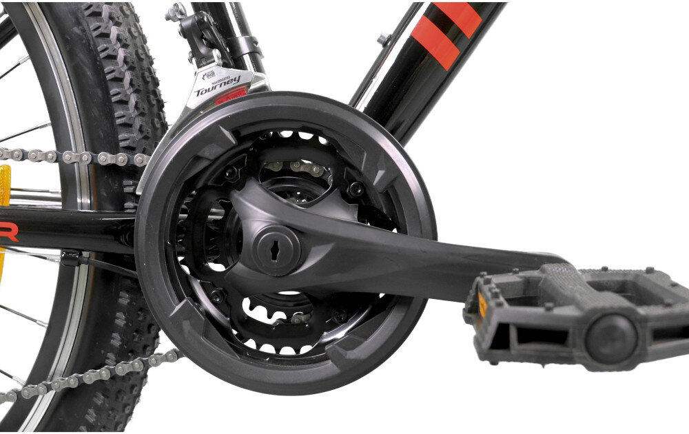 Zbliżenie na mechanizm korbowy roweru z czarną osłoną i pedałem. Widoczny jest fragment opony z bieżnikiem oraz napis \'Tourney\' na przerzutce.