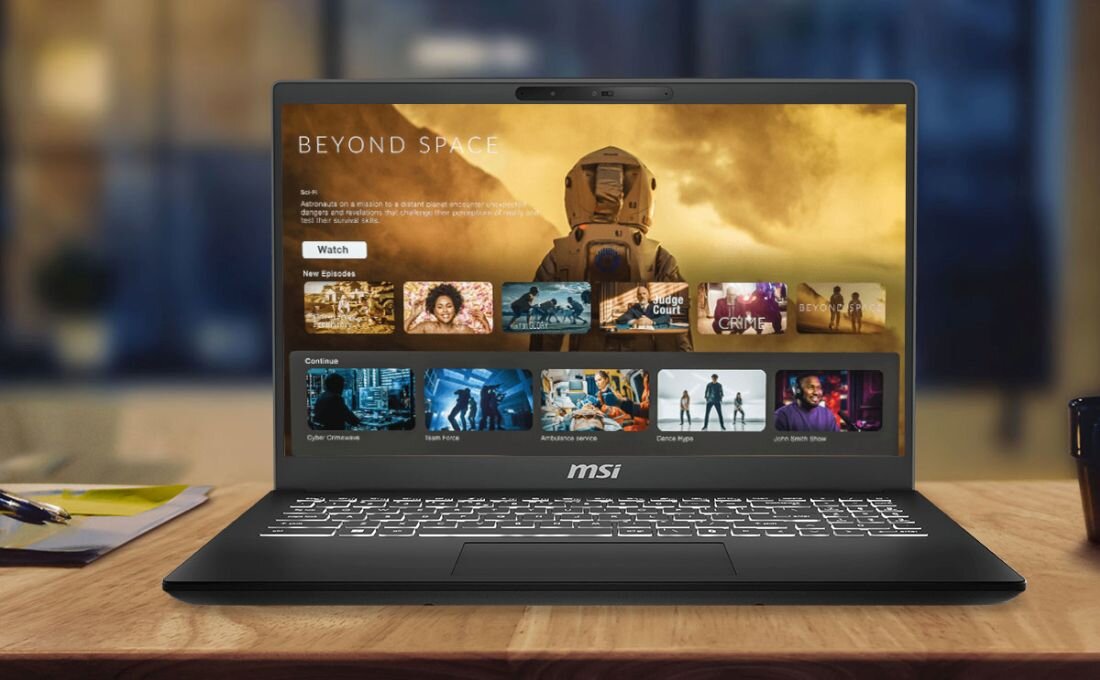 Laptop MSI Modern 15 H ustawiony na drewnianym biurku, ekran prezentuje interfejs platformy streamingowej z serialem science fiction „Beyond Space”, podświetlana klawiatura, rozmyte tło biurowe z kubkiem po prawej stronie. - Silnik MSI AI Engine   