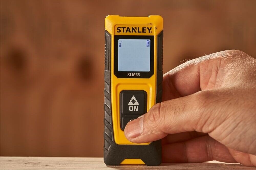 Dalmierz laserowy STANLEY SLM65 STHT77065-0 20m, odległości, meble, pamięć, IP40, kciuk dłoni wciska przycisk na stojącym na blacie dalmierzu