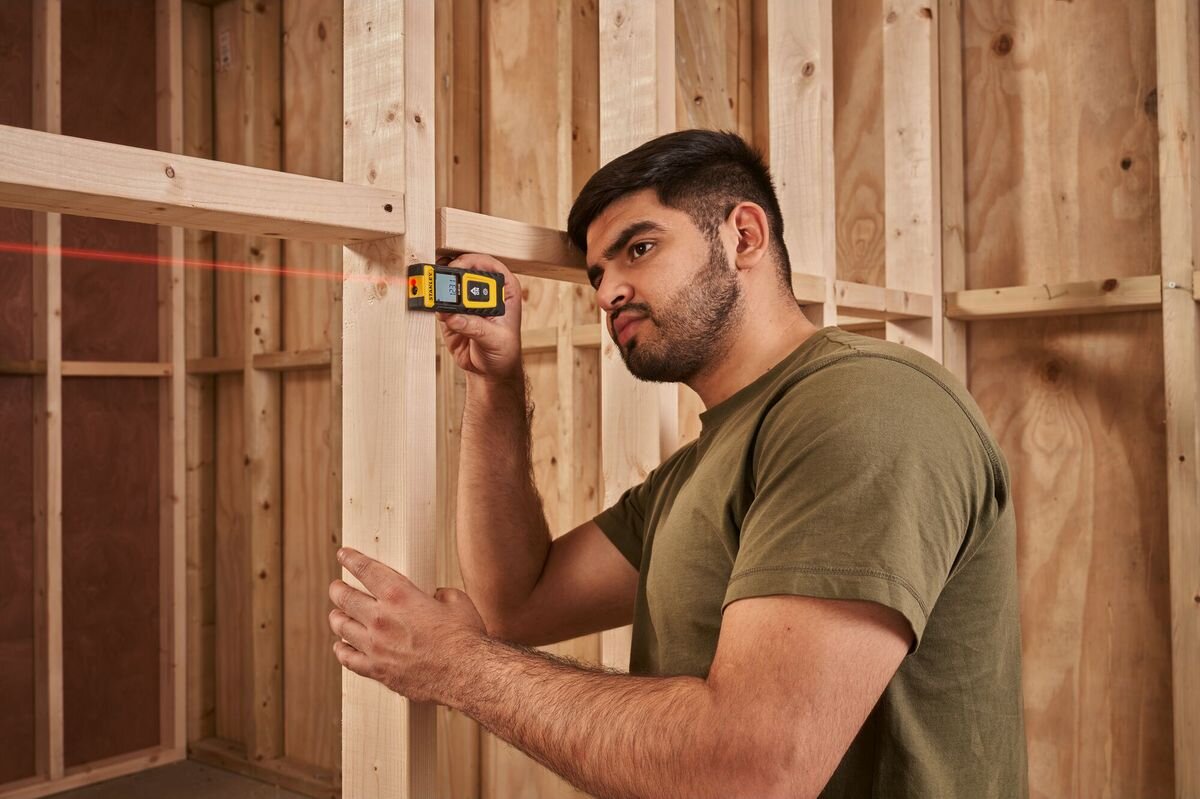 Dalmierz laserowy STANLEY SLM100 STHT77100-0 30m, laser, odległość, powierzchnia, dokładność, mężczyzna odmierza laserem dystans w pomieszczeniu