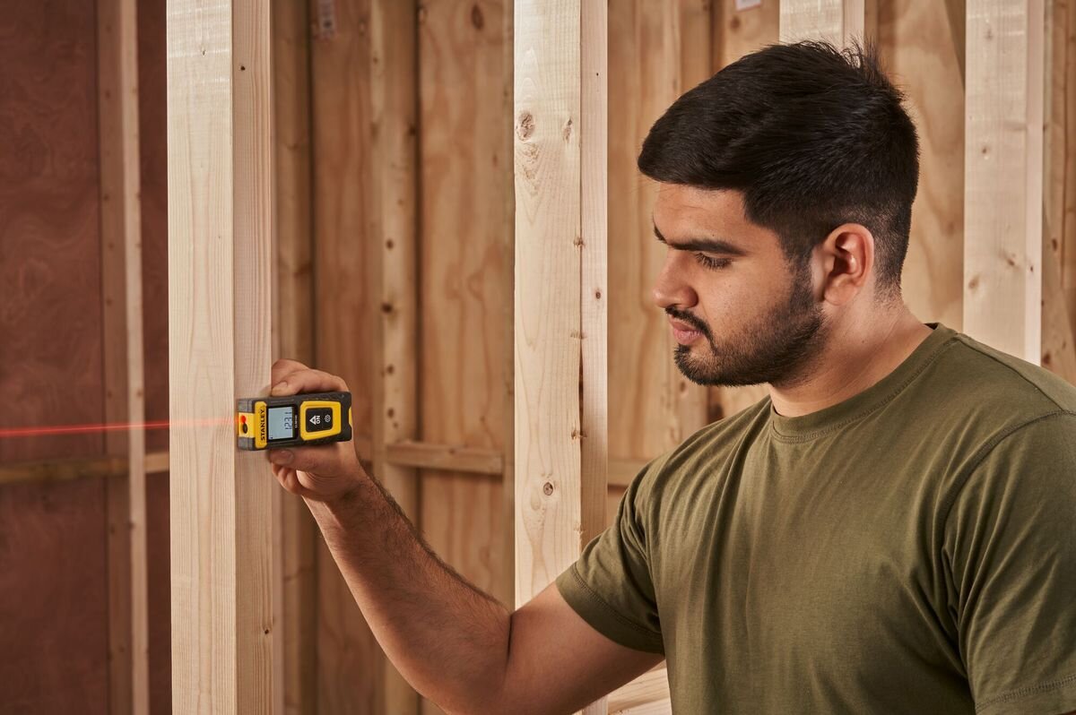 Dalmierz laserowy STANLEY SLM100 STHT77100-0 30m, dwa przyciski, intuicja, ergonomia, ciasne, mężczyzna odmierza laserem dystans w pomieszczeniu
