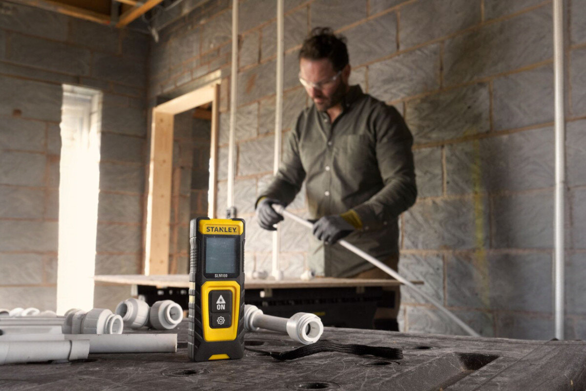 Dalmierz laserowy STANLEY SLM100 STHT77100-0 30m, 0.15-30m, remonty, materiały, majsterkowanie, mężczyzna pracuje przy stole warsztatowym, na pierwszym planie dalmierz