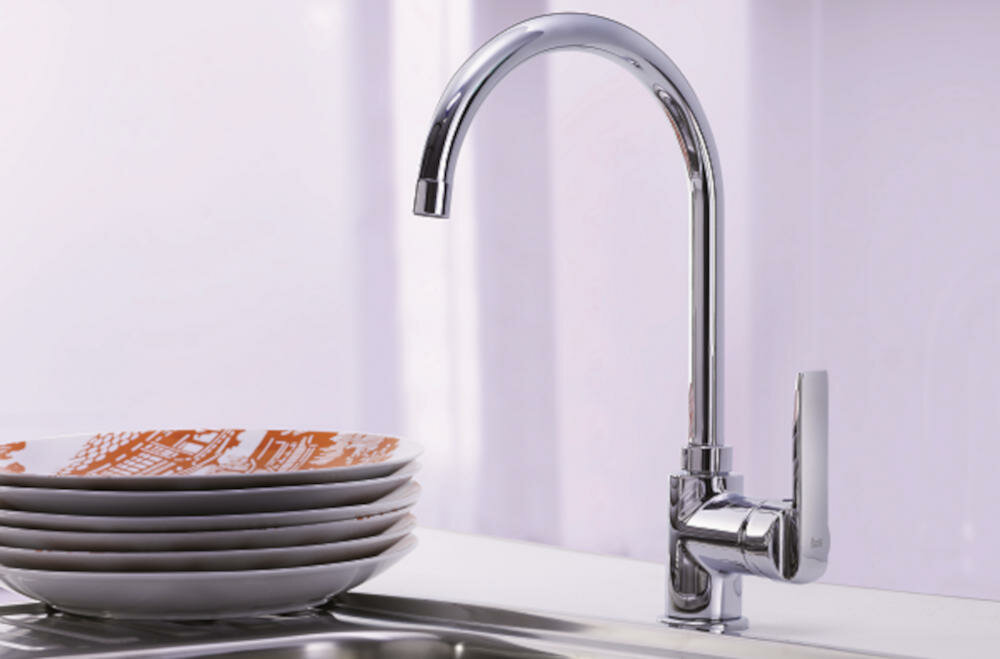 Na obrazie widoczna jest chromowana bateria kuchenna z wysokim, zakrzywionym wylewem. Obok znajduje się stos talerzy z pomarańczowym wzorem na białym tle.