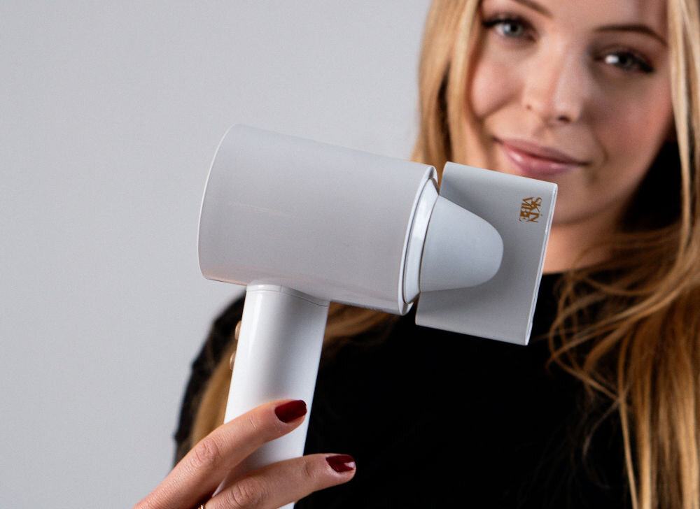 Suszarka SKINVIBE Libra Airloom SV-002WH Biały 1500W osoba trzyma w ręce biała suszarkę przyczepiona końcówka szare tło system jonizacji naturalna harmonia miękkie w dotyku włosy jedwabisty blask sprężystość wymagające warunki funkcjonalność troska o piękno olśniewający wygląd