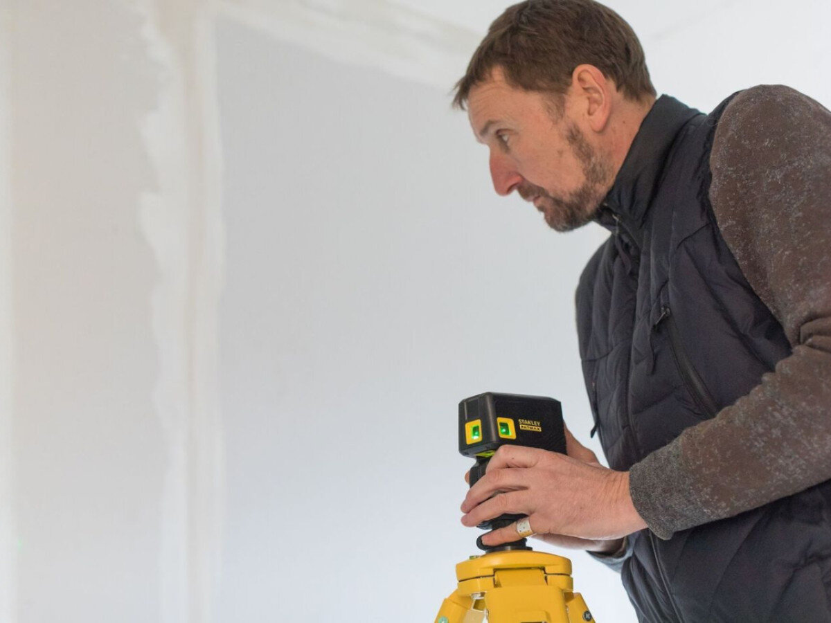 Laser krzyżowy STANLEY Fatmax FMHT77596-1, skos, schody, ramię, blokada, mężczyzna stoi z laserem i tripodem przed ścianą