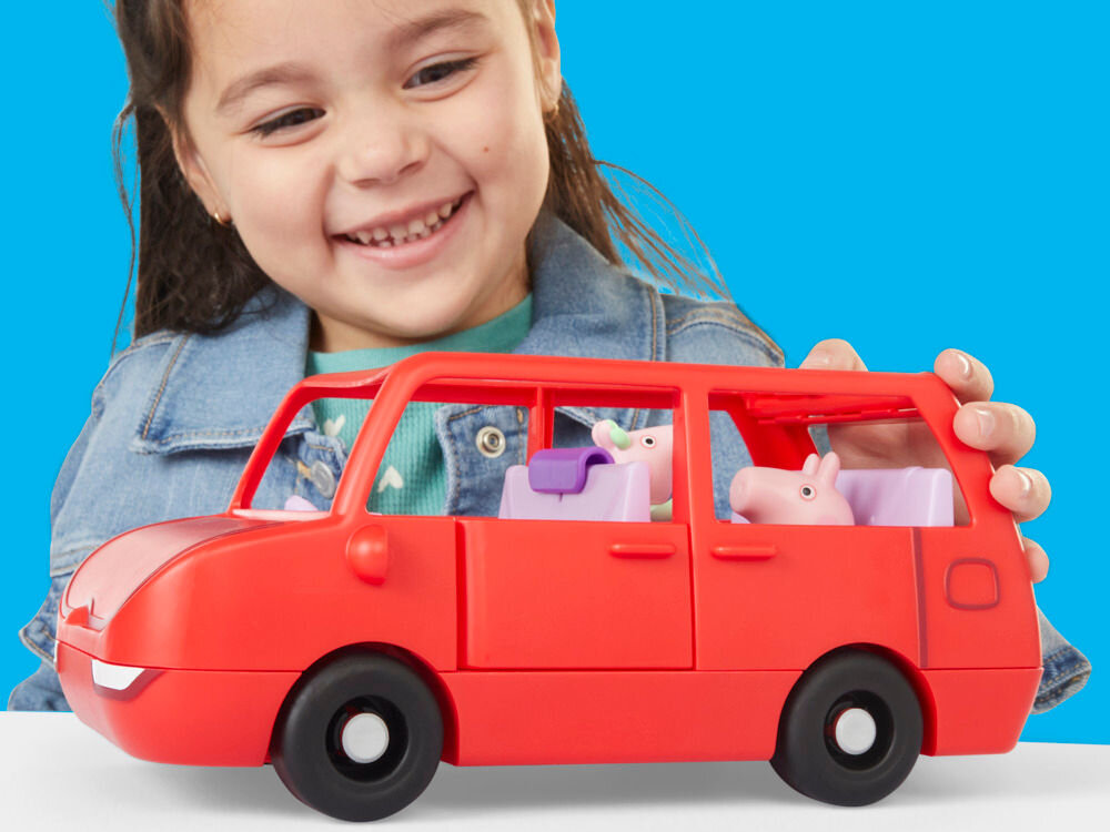 Zestaw figurek HASBRO Świnka Peppa Duży pojazd G05415L0 Zdjęcie z góry na czerwonego zabawkowego vana Peppa Pig. Jedna ręka dziecka podnosi fioletowe siedzenie kierowcy, a druga trzyma dach. Bagażnik auta jest otwarty i widać w nim błękitną torbę. W tylnym rzędzie siedzą figurki George'a i Peppy. Tło błękitne.
