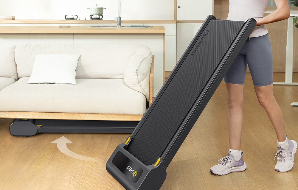 Bieżnia elektryczna UREVO E3S 1654.87W 6.4 km-h Kobieta łatwo wsuwająca płaską bieżnię pod sofę w salonie, prezentująca oszczędność miejsca wbudowane miejsce na pilota możliwość szybkiego złożenia wymiary po złożeniu 127 x 53 x 12 cm