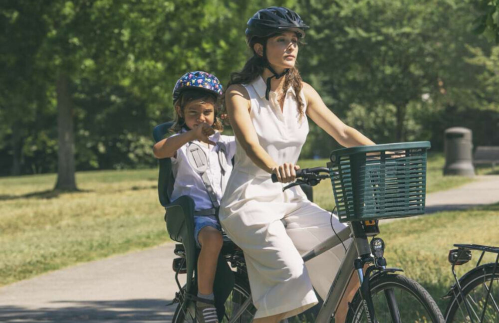 Fotelik rowerowy BELLELLI Mr Fox Clamp Czarny park osoba w kasku jedzie na rowerze na bagażniku z tyłu siedzi dziecko w foteliku w tle drzewa droga bezpieczna podróż z dzieckiem komfort wygoda certyfikaty