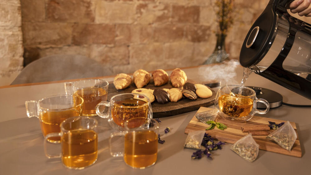 Czajnik CECOTEC ThermoSense 330 Glassy osoba trzyma czarny czajnik nalewa wodę do filiżanki z herbatą obok stoi kilka filiżanek z jasnym napojem 26,9 x 22,5 x 18,2 cm pojemność 1,7 litra dla większych rodzin miłośników częstych spotkań przy herbacie do 10 filiżanek napoju za jednym razem szybkość wygoda serwowanie ulubionych naparów wszystkim domownikom jednocześnie