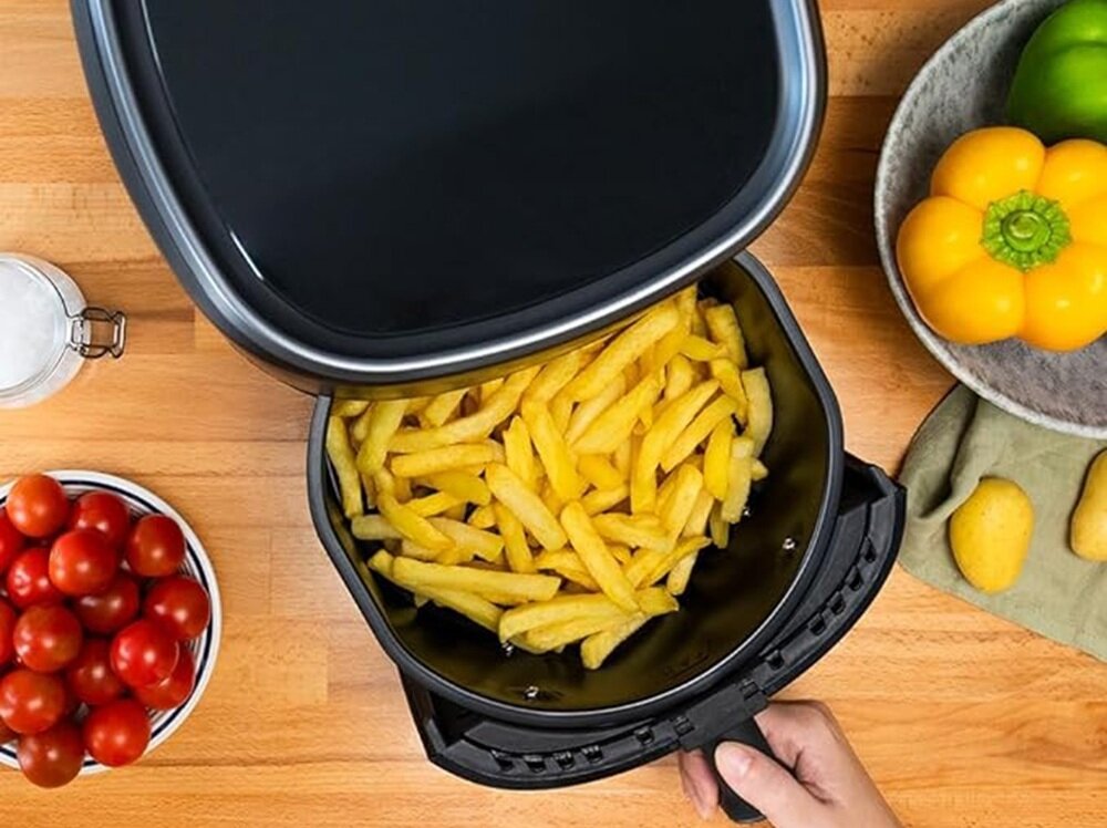 Na drewnianym blacie stoi otwarta frytkownica z frytkami w środku. Obok znajdują się miski z pomidorami i papryką oraz kilka ziemniaków.