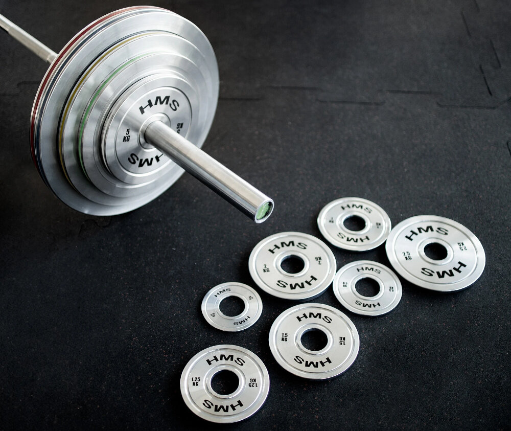 Obciążenie HMS TOS150 (15 kg) siłownia ciemna podłoga gryf na gryfie założone obciążenia przed gryfem leży kilka obciążeń precyzja trening siły budowanie formy zdrowie