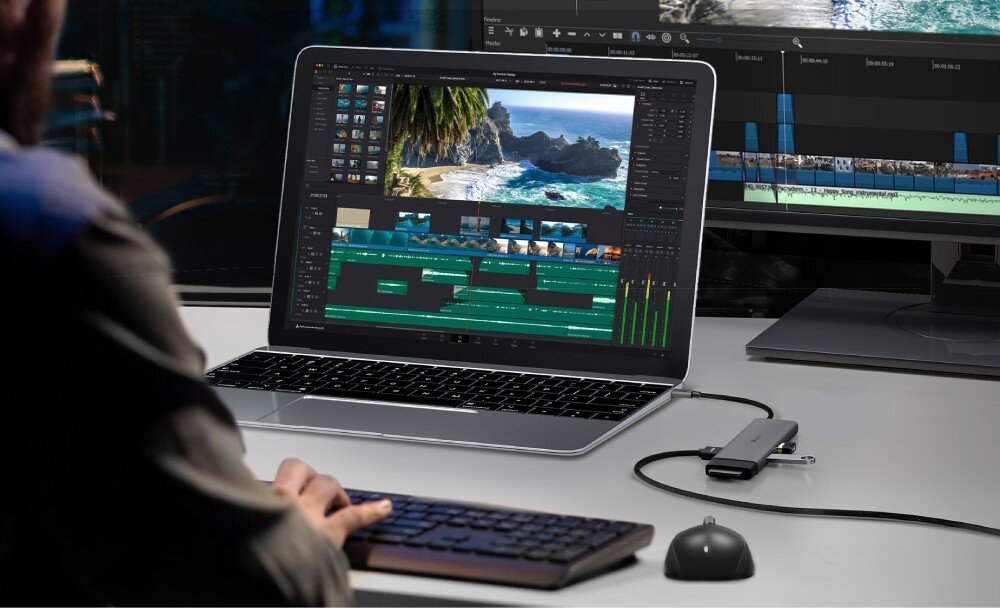 Hub TP-LINK UH7020C Osoba siedząca przy biurku pracuje na laptopie z otwartym programem do edycji wideo, do którego podłączony jest szary adapter z wieloma kablami i akcesoriami, a obok znajduje się drugi monitor wyświetlający oś czasu projektu
