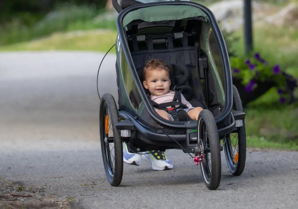 Na obrazie widoczna jest przyczepka rowerowa z dzieckiem w środku, poruszająca się po ścieżce. Przyczepka ma zielono-czarny kolor i jest wyposażona w pasy bezpieczeństwa.