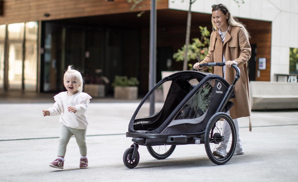 Przyczepka rowerowa HAMAX Cocoon One Szaro-czarny miasto osoba prowadzi przed sobą przyczepkę przed nimi idzie dziecko wycieczka rowerowa podróż