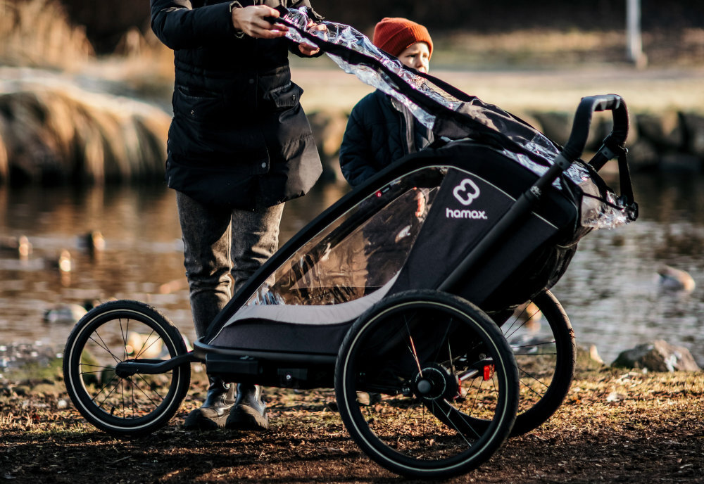 Przyczepka rowerowa HAMAX Cocoon One Szaro-czarny jezienna sceneria osoba zaklada osłonę na przyczepkę obok stoi dziecko Zintegrowana osłona przeciwdeszczowa Regulowana osłona przeciwsłoneczna górne okienko