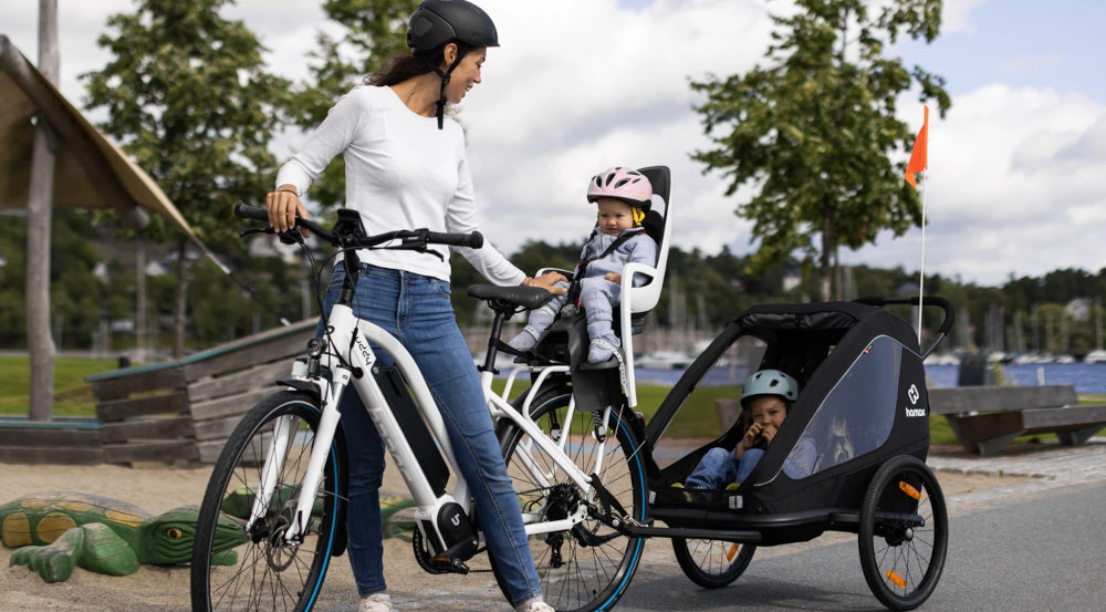Przyczepka rowerowa HAMAX Traveller Twin Czarny ulica osoba z dzieckiem w foteliku obraca się na dziecko siedzące w przyczepce wycieczka rowerowa przewożenie dzieci bezpieczeństwo wygoda