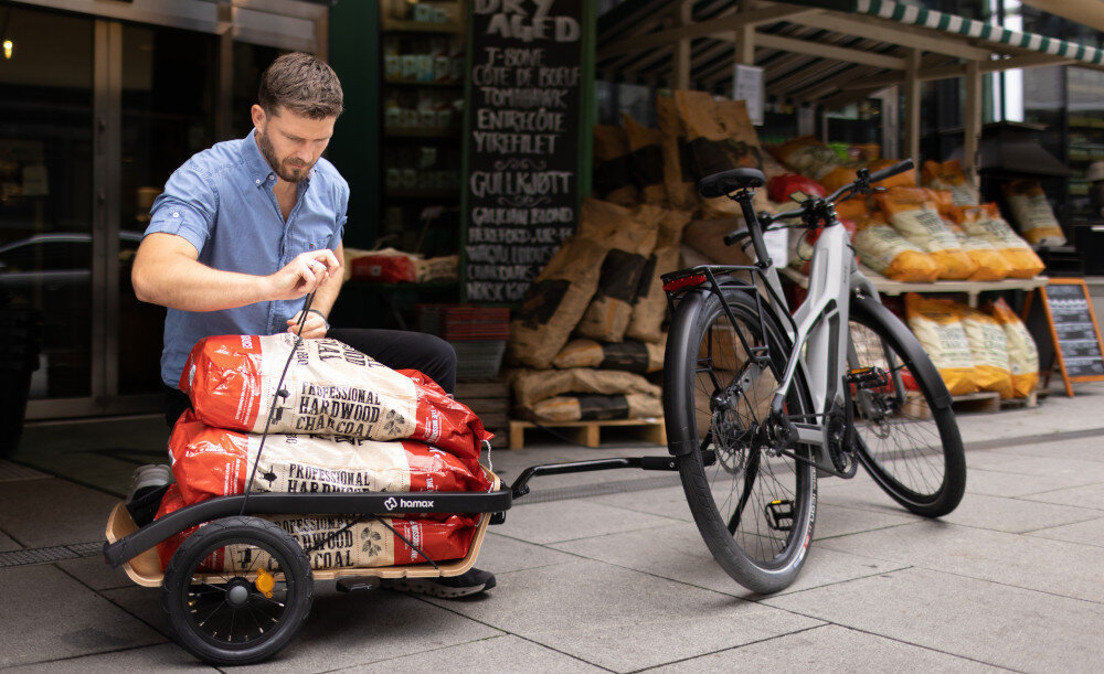 Przyczepka rowerowa HAMAX Venture+ 400702 osoba klęczy przy przyczepce przed sklepem zabezpiecza ładunek na przyczepce Platforma ładunkowa Aluminiowa rama wewnątrz uniwersalne rowki Maksymalne obciążenie szyn wynosi do 15 kg