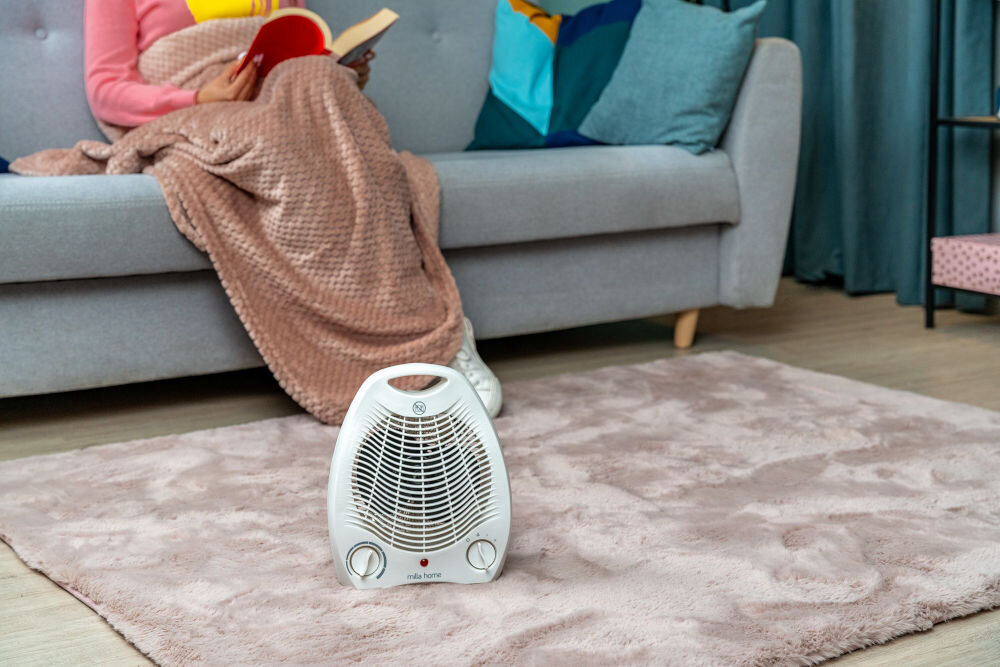 Termowentylator MILLA HOME MFH103 Biały termowentylator MILLA HOME MFH103 stojący na puszystym dywanie w salonie obok osoby czytającej książkę na sofie.