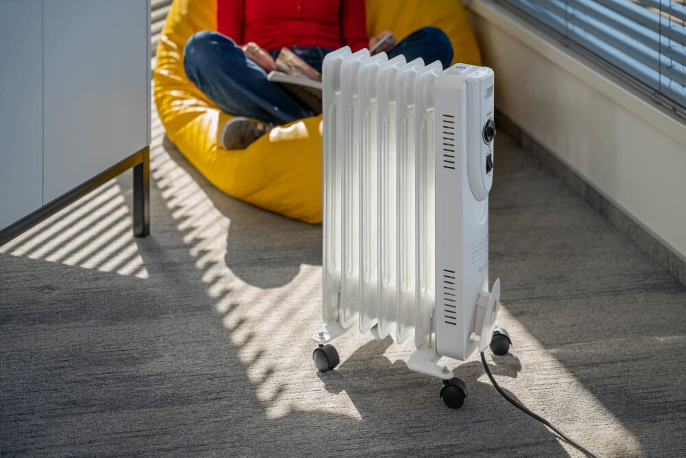 Grzejnik olejowy MILLA HOME OH702 1500W Biały, żebrowy grzejnik olejowy na kółkach stojący w pokoju. W tle na żółtym pufie siedzi osoba czytająca książkę. Przez żaluzje w oknie wpadają paski światła