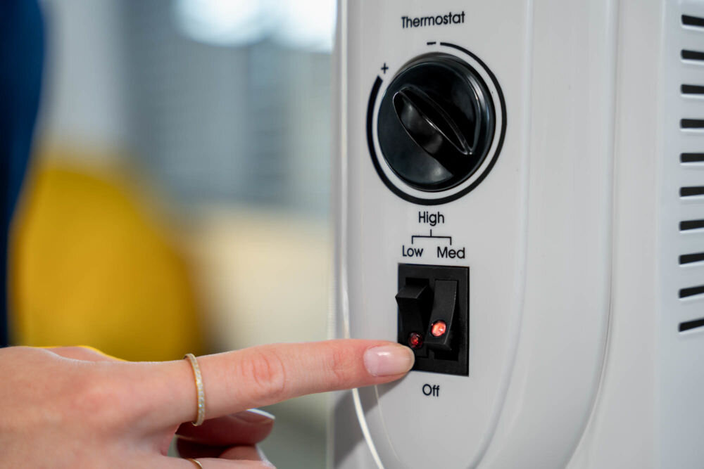 Grzejnik olejowy MILLA HOME OH702 1500W z trzystopniową regulacją mocy grzania do efektywnego ogrzewania - Zbliżenie na panel sterowania białego grzejnika olejowego. Palec użytkownika włącza przełącznik zasilania (świecący na czerwono) obok oznaczeń High, Low, Med i pokrętła termostatu