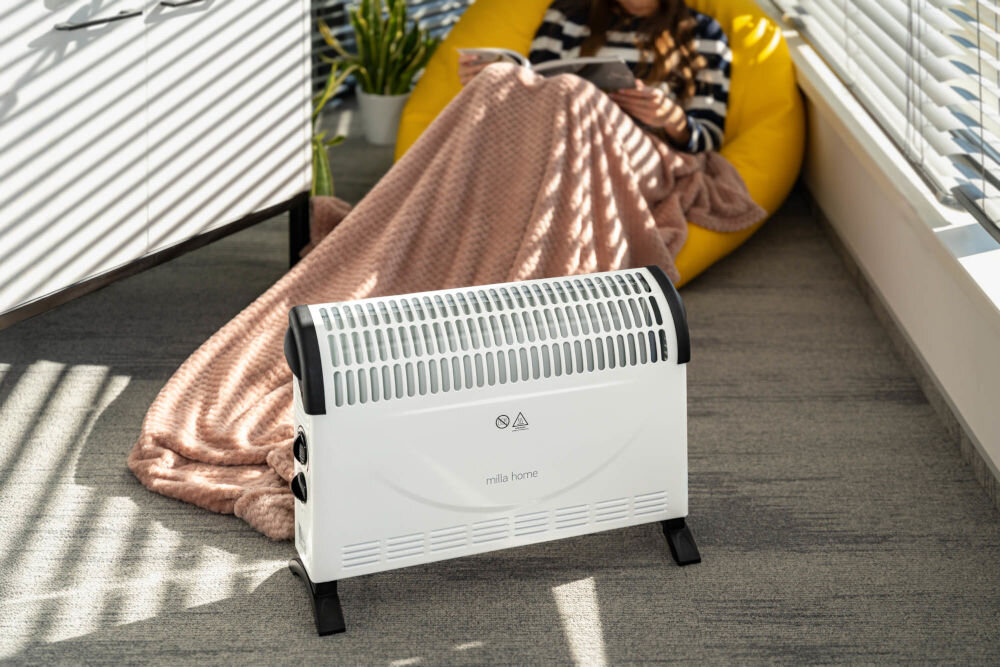 Grzejnik konwektorowy MILLA HOME MCH103 stojący w pokoju w tle osoba nakryta kocem siedząca na fotelu