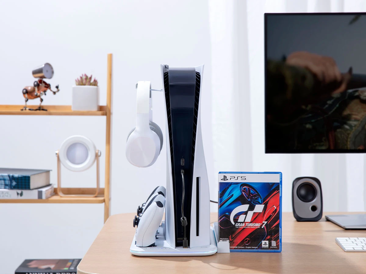 Na biurku znajduje się konsola PlayStation 5 z podstawką, na której umieszczone są dwa kontrolery i słuchawki. Obok konsoli stoi pudełko z grą \'Gran Turismo 7\'.