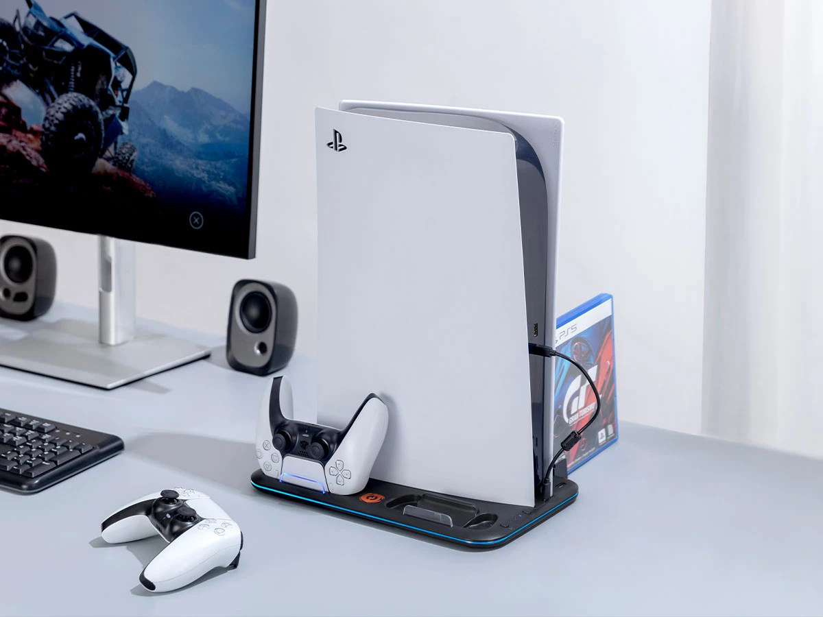 Na biurku znajduje się konsola z logo PlayStation, umieszczona na podstawce z kontrolerem. Obok konsoli widoczna jest okładka gry PS5 oraz monitor z obrazem terenowego pojazdu.