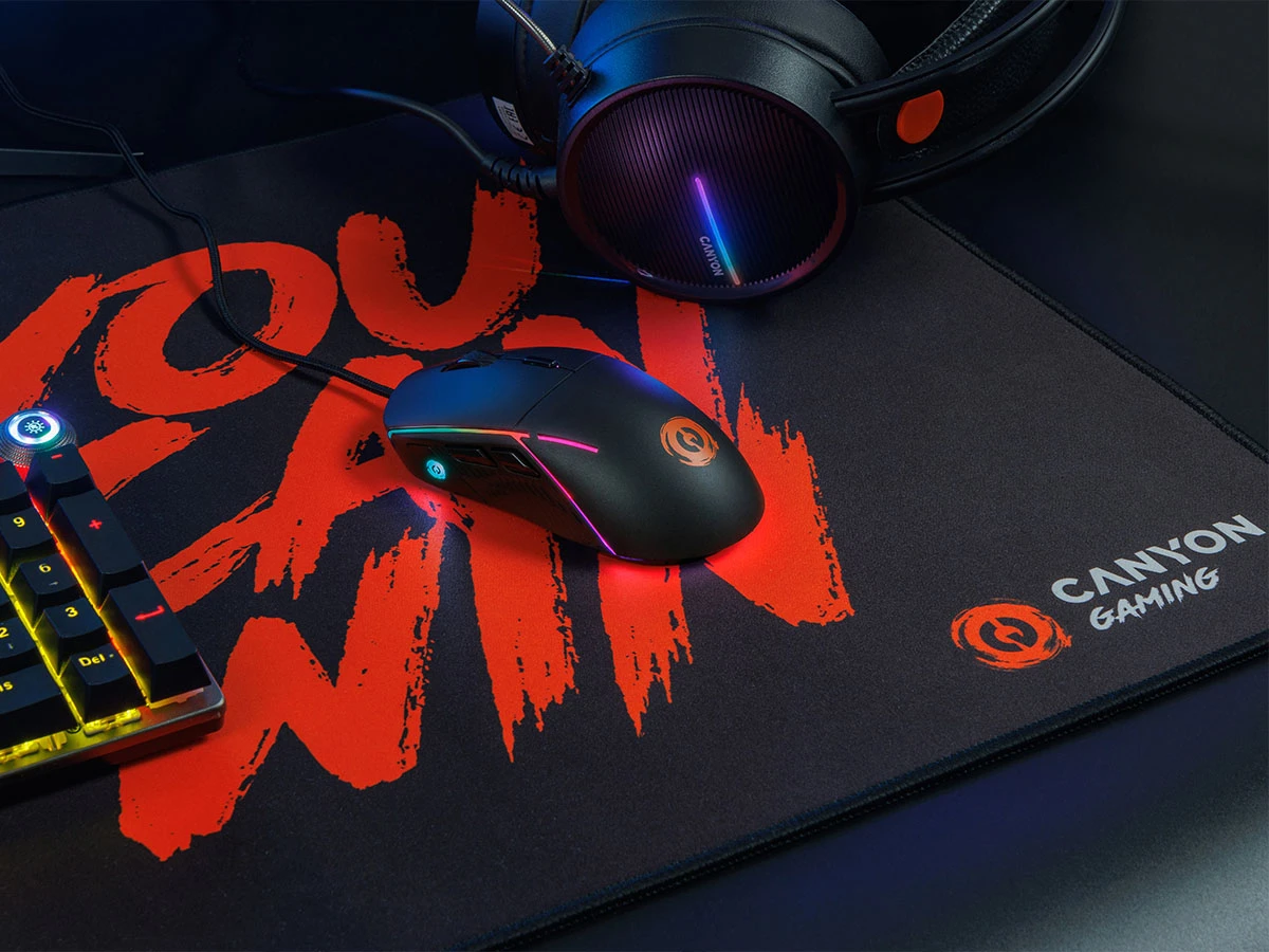 Na obrazie znajduje się czarna mysz gamingowa z podświetleniem RGB, leżąca na podkładce z napisem \'YOU CAN WIN\'. Obok widoczne są słuchawki oraz klawiatura, a na podkładce znajduje się logo \'CANYON GAMING\'.