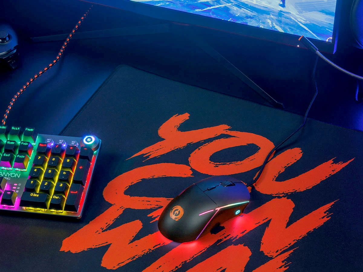 Na obrazie znajduje się czarna mysz gamingowa z podświetleniem RGB, umieszczona na podkładce z napisem \'YOU CAN WIN\'. Obok widoczna jest klawiatura z kolorowym podświetleniem oraz logo \'CANYON GAMING\'.