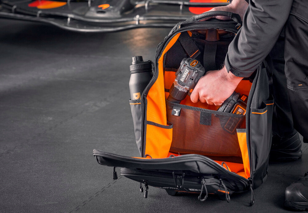 Plecak monterski DEWALT McLaren F1 DWST60101-9 osoba wkłada wiertarkę do plecaka widoczny bidon i siatkowa kieszeń główna komora otwiera się szeroko szybki dostęp do zawartości jasna żółta tkanina wyściełająca wnętrze dobra widoczność narzędzi zamek błyskawiczny z funkcją szybkiego zamykania
