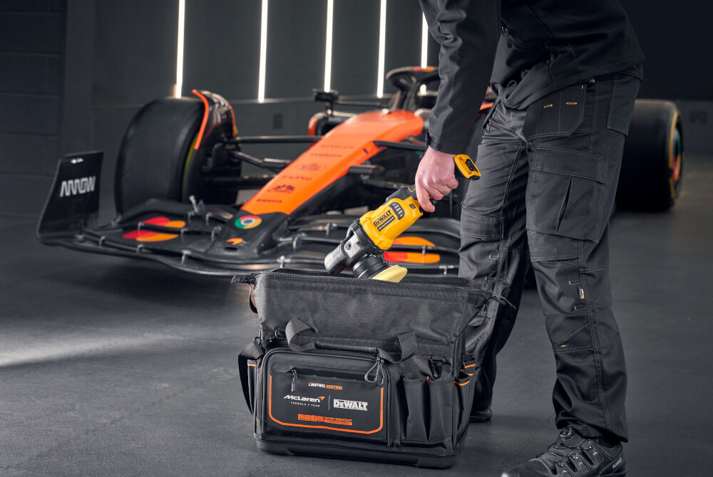 Torba narzędziowa DEWALT DWST60104-9 osoba wkłada narzędzie do torby w tle samochód McLaren główna kieszeń otwiera się szeroko składana konstrukcja łatwy dostęp do wszystkich przegródek jasna żółta tkanina wewnątrz zamek błyskawiczny z funkcją szybkiego zamykania powiększony schowek na dłuższe narzędzia kieszeń na butelkę osobne komory na wiertarkę i akumulatory