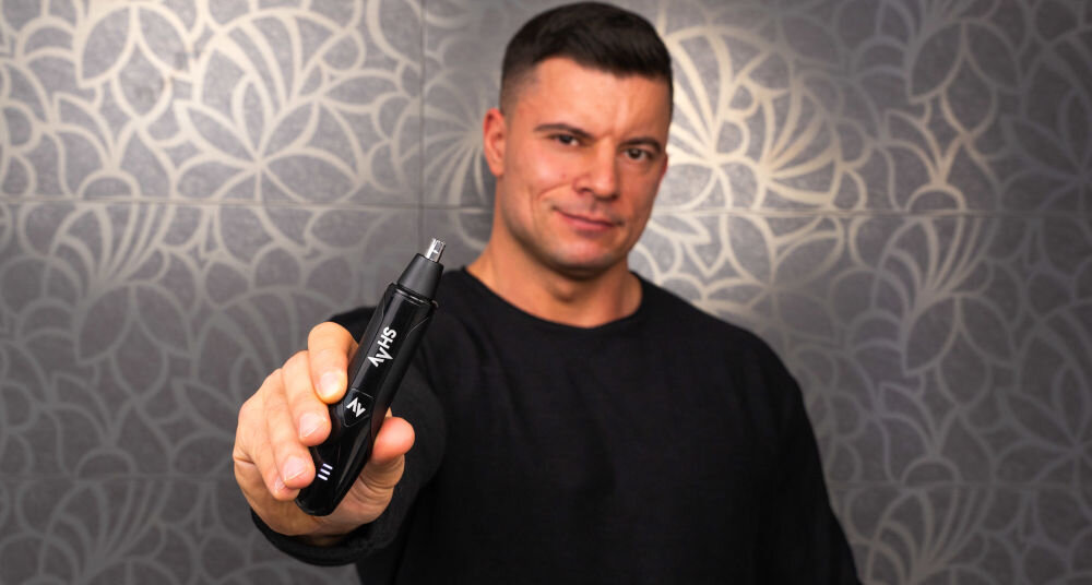 Trymer SHAV Nose Trimmer optymalna gładkość mężczyzna trzyma w ręku czarny trymer uśmiecha się ciemne tło