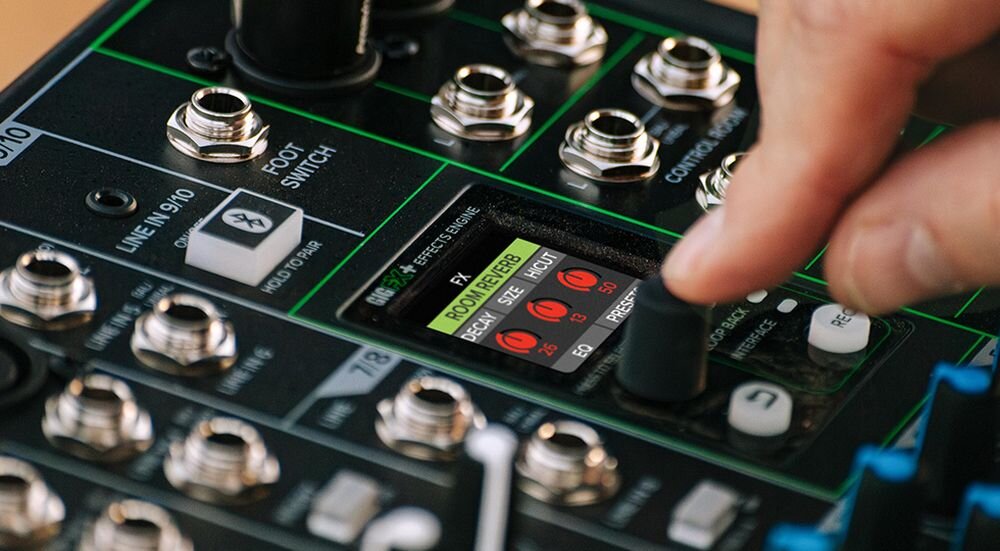 Mikser audio MACKIE ProFX10 GO efekty GigFX+ reverb delay chorus wyświetlacz LCD, ręka trzymająca guzik, mikser stojący na stole