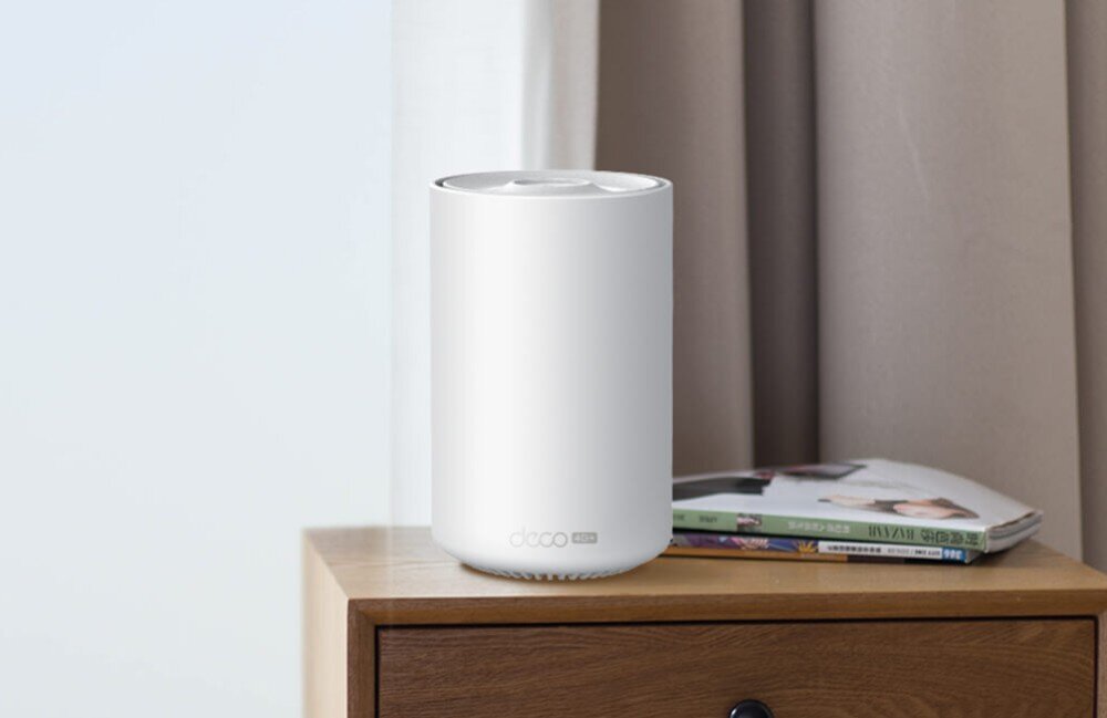 Router TP-LINK Deco X10-4G Biały, cylindryczny router stojący na drewnianej szafce obok czasopism w jasnym pomieszczeniu z zasłoną w tle, opis