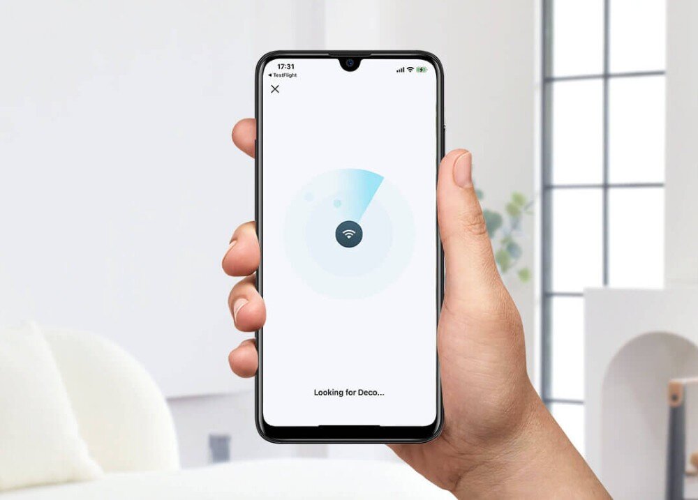 Router TP-LINK Deco X10-4G Dłoń trzymająca smartfon z uruchomioną aplikacją wyszukującą urządzenie sieciowe, w tle jasne wnętrze salonu, aplikacja, funkcje, narzędzia