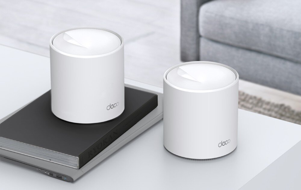 Router TP-LINK Deco X50 Pro Dwa białe cylindryczne urządzenia stojące na stole obok siebie, jedno z nich umieszczone na grubej książce z napisem 'Ryokan', opis