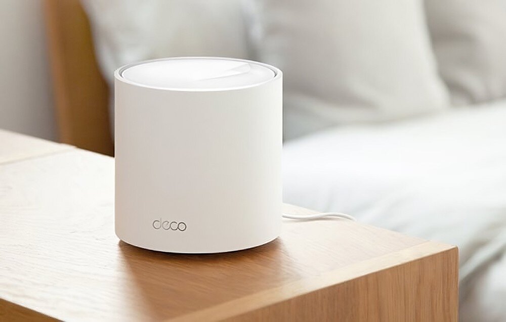 Router TP-LINK Deco X50 Pro Jedno białe cylindryczne urządzenie stojące na drewnianym stoliku obok łóżka z pościelą w tle, sterowanie, komendy głosowe