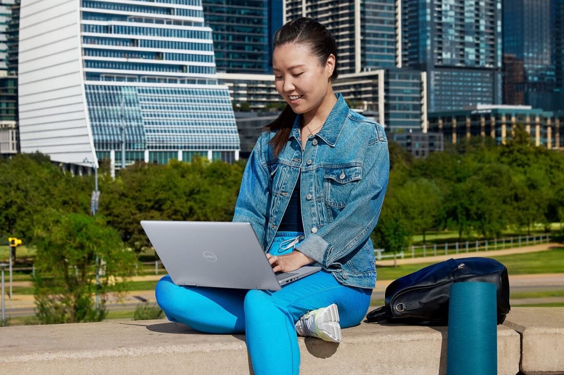 Kobieta pracuje na laptopie, siedząc na ławce w parku, w otoczeniu drzew i krzewów. - Niska waga i kompaktowy rozmiar 