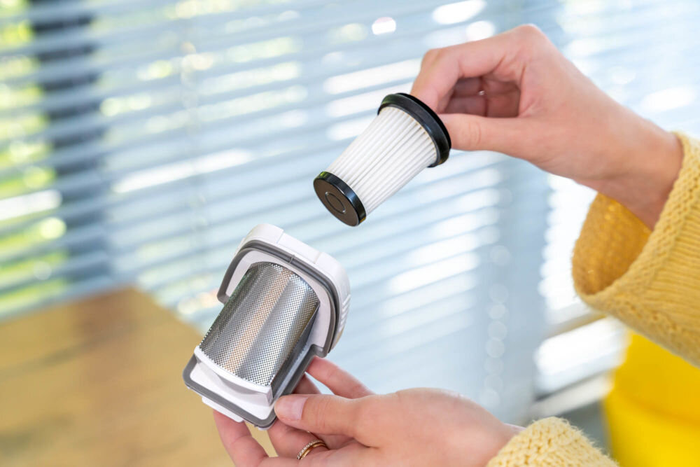 Filtr do odkurzacza GÖTZE & JENSEN HV800 HVA800MF (3 sztuki) Dłonie prezentują zestaw filtrów: jedna trzyma szarą obudowę z metalowym filtrem wstępnym, a druga trzyma biały, plisowany filtr EPA gotowy do montażu w obudowie. Tło stanowią okienne żaluzje.