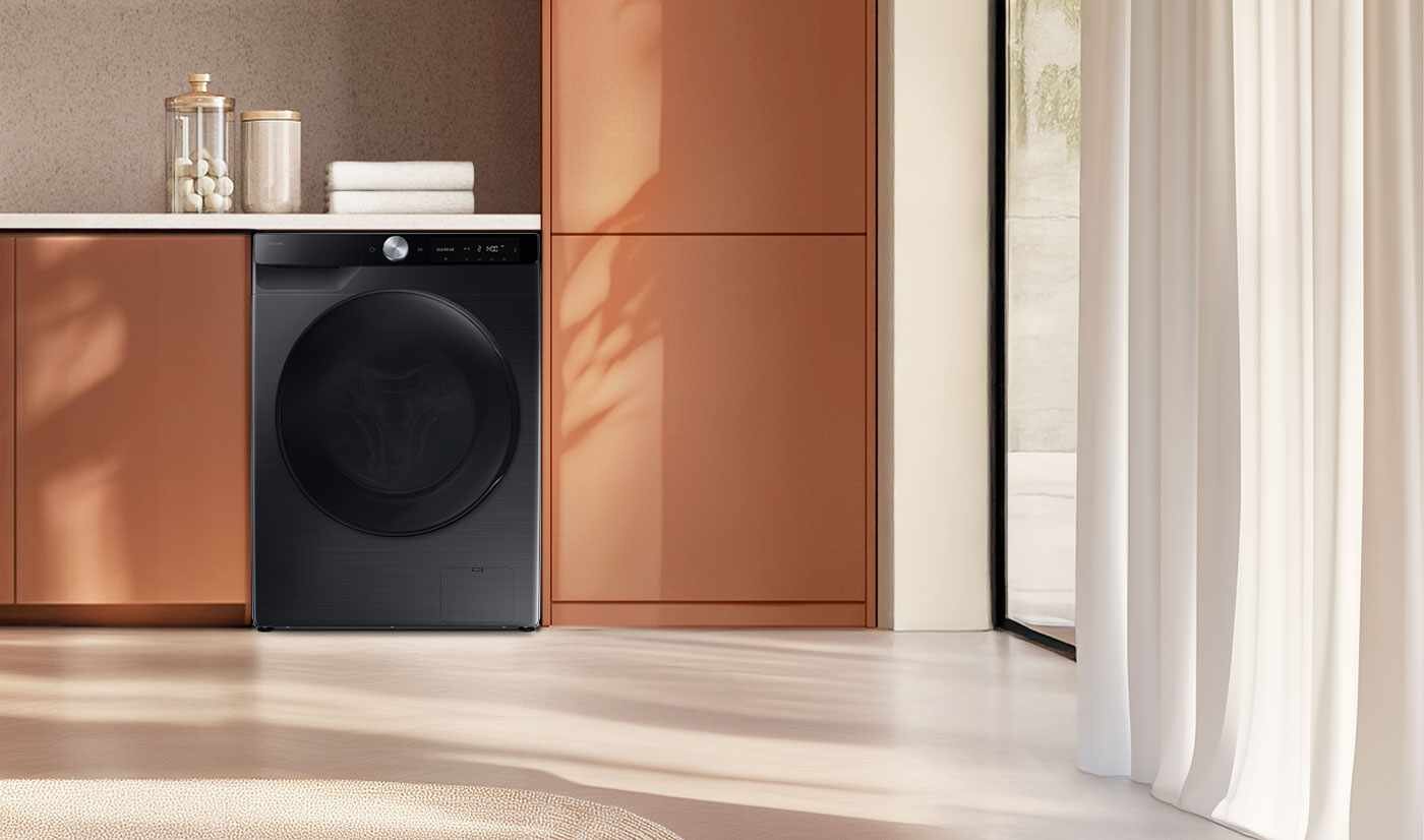 Pralko-suszarka Samsung WD90DG6G94BBU4 9 kg - grafika główna Czarna pralko-suszarka w nowoczesnej kuchni, obok szafek w kolorze miedzianym. Na blacie znajdują się złożone ręczniki i szklany pojemnik z pokrywką.