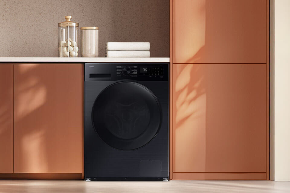 Lodówka Samsung Side by Side RS80F66KCFEO Czarna pralko-suszarka w zabudowie kuchennej, obok której znajdują się szafki w kolorze brązowym. Na blacie stoją dwa słoiki i złożone ręczniki.