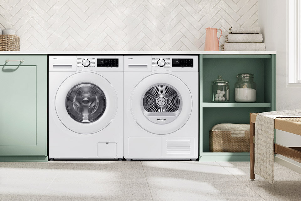 Na obrazie znajdują się dwie białe pralki ustawione obok siebie w nowoczesnej kuchni. Obok nich znajdują się półki z dekoracjami i ręcznikami.