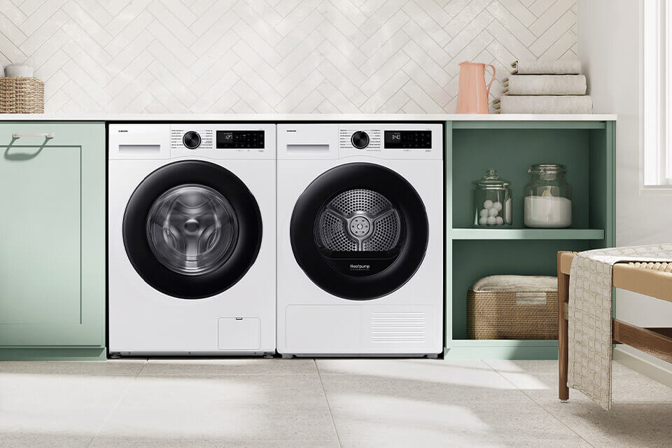 Na obrazie znajdują się dwie białe pralki z czarnymi drzwiami, umieszczone obok siebie w jasnej kuchni. Obok pralek znajdują się półki z dekoracjami i koszami.