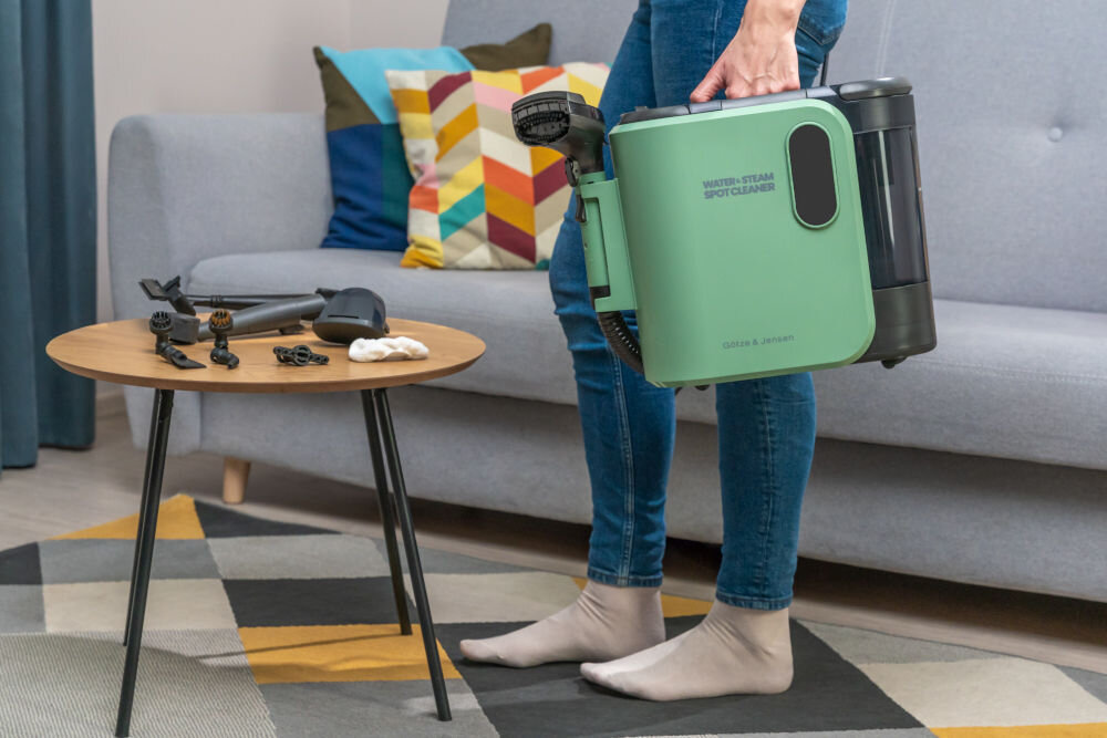 ODKURZACZ PIORĄCY SPOTCLEAN GOTZE&JENSEN CSC600 Kobieta w dżinsach i skarpetkach trzyma podniesiony zielono-czarny odkurzacz piorący za uchwyt. Na stoliku obok leżą akcesoria.